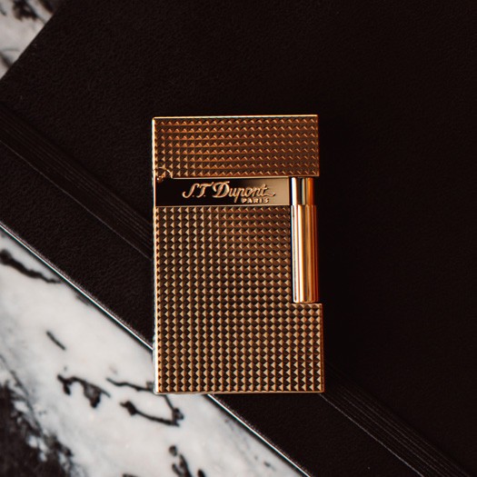 S.T. Dupont Ligne 2 Yellow Gold (16284 / REF. 016284) е емблематична луксозна запалка, съчетаваща масивен месингов корпус, финиш в жълто злато и прецизна Diamond Head декорация. Множеството геометрични фасети улавят и пречупват светлината, придавайки на златистата повърхност характерен динамичен блясък. Моделът е оборудван с двоен мек пламък и носи характерното акустично усещане при отваряне, с което Ligne 2 е известна сред ценителите. С класически размери 37 × 62 × 11 mm, 16284 съчетава солидно присъствие, прецизна механика и неподвластна на времето елегантност – истинска златна класика в света на S.T. Dupont.